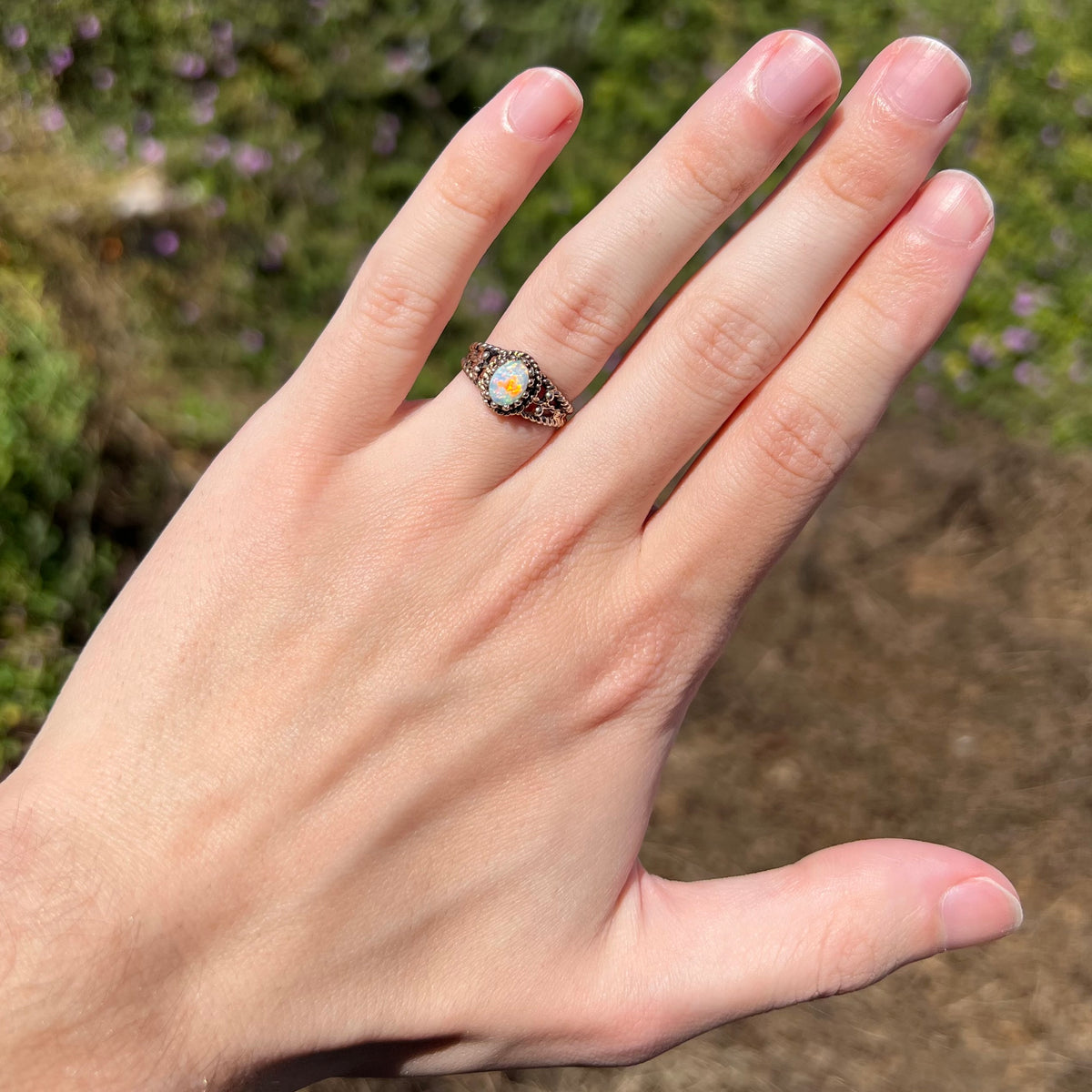 Hand wearing a used, estate Australian opal in brass ring under natural sunilght; the opal has a bright red flash of color, seen from a distance