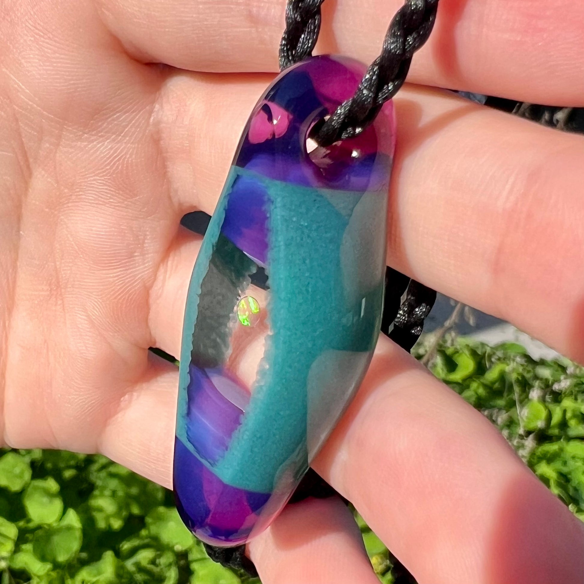 An opal in glass pendant by Utah artist, Nathan Snyder, shown from an angle in the sunlight while held in a hand.  The opal has green colors.