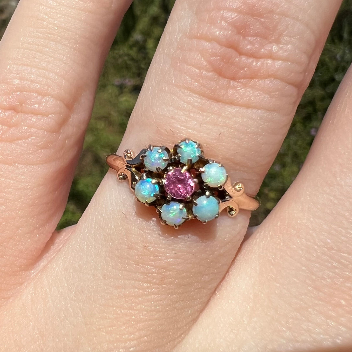 Close-up of 9k gold Victorian ring set with six round Hungarian opals and a pink tourmaline center stone, worn on finger under sunlight