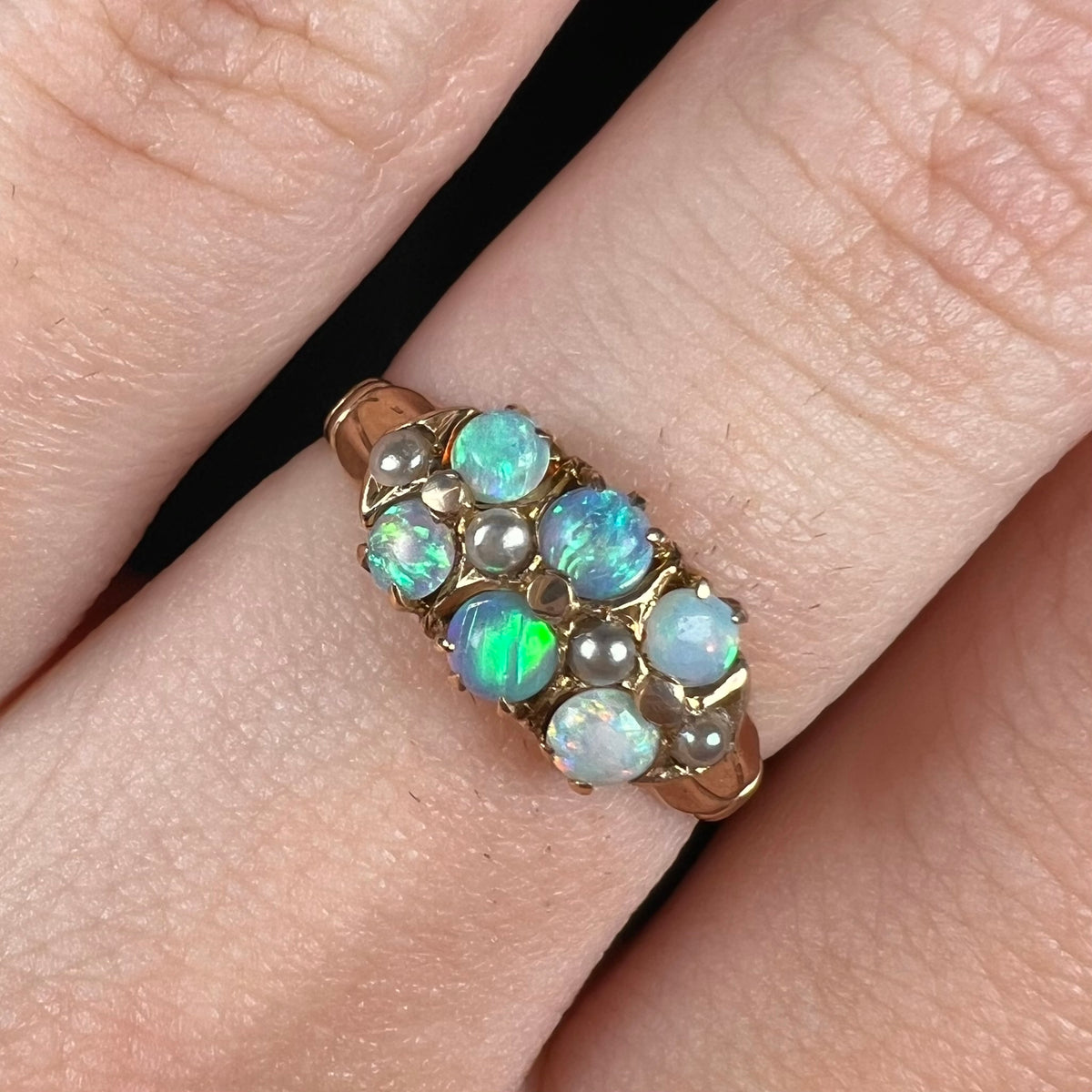 Close-up macro view of Victorian six-stone opal and pearl-accented ring, worn on finger; the opals shine with green and blue colors