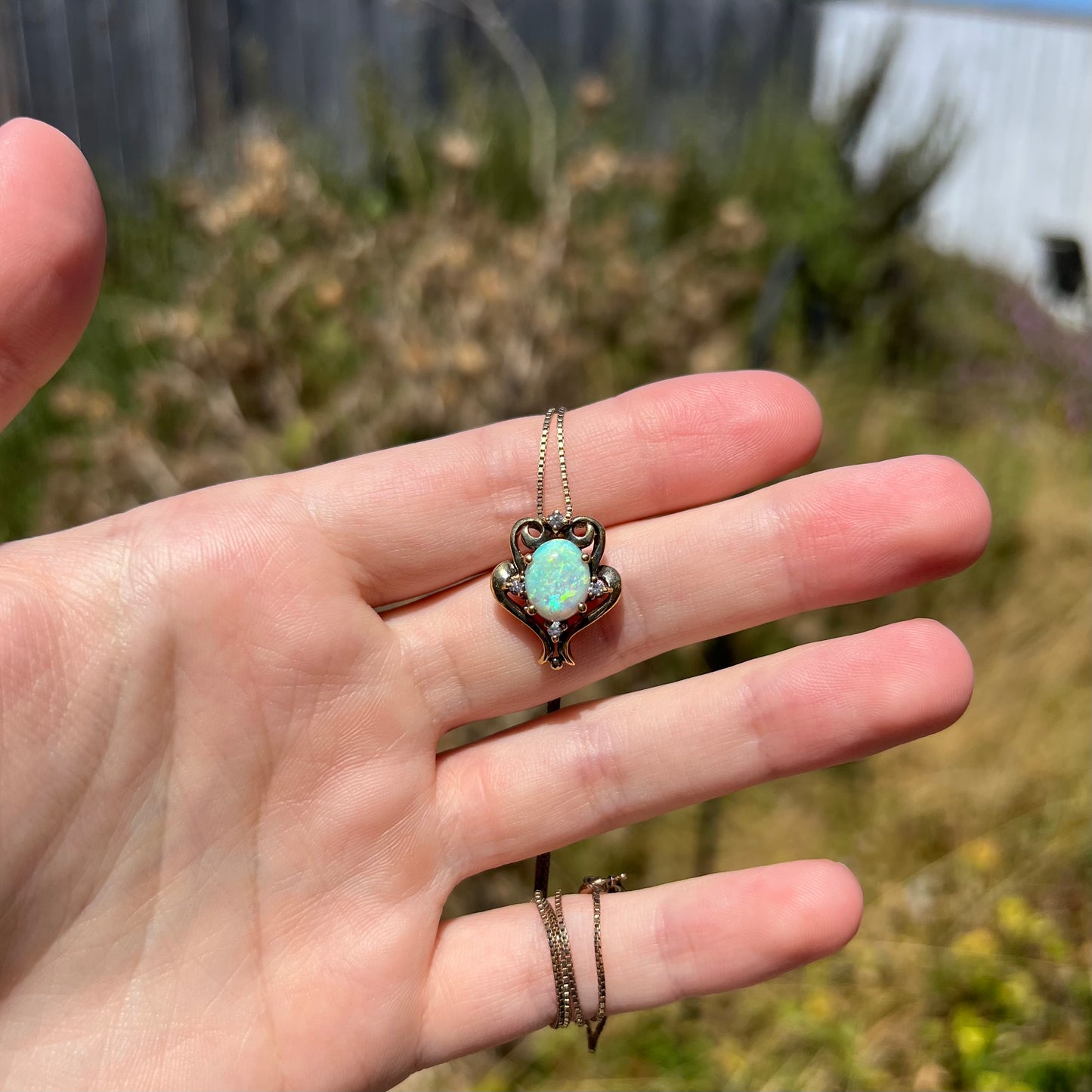 Hand holding a women's Victorian-style oval-shaped opal and CZ necklace in sunlight against a blurred outdoor background.