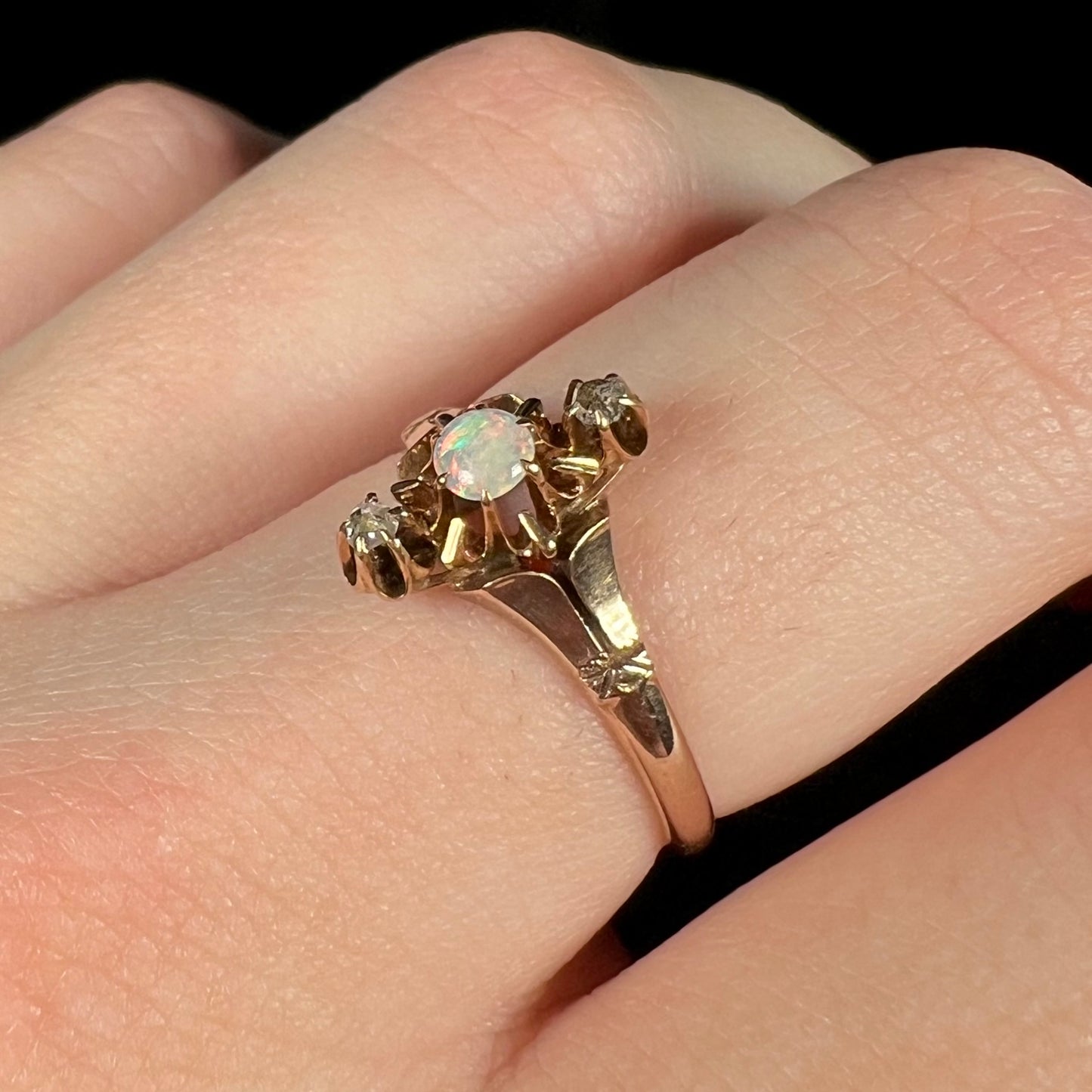 Angled close-up of a Victorian prong-set three-stone opal and diamond ring worn on a finger against a black background.