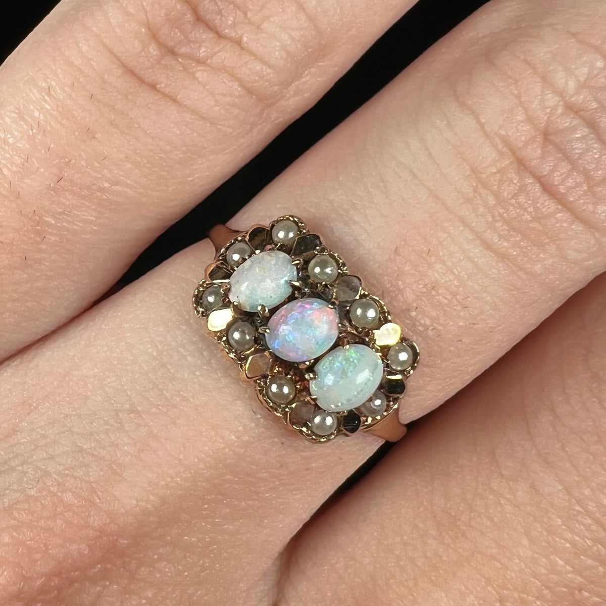 Close-up of three-stone Victorian ring set with White Cliffs opals and a halo of seed pearl accents in 9k gold, worn on a finger