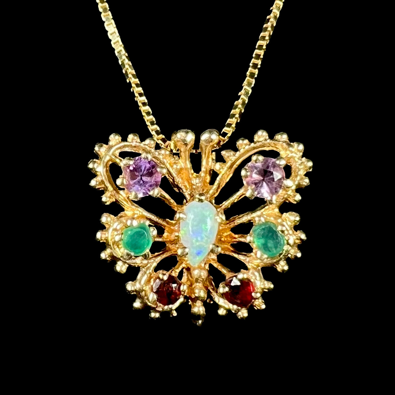 Close-up macro of a vintage 14k gold opal, amethyst, emerald, and garnet butterfly pendant shown from front against black background.