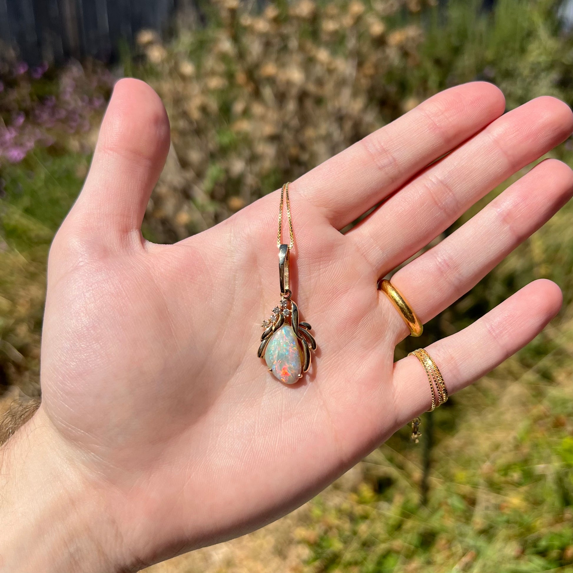 A vintage, 1980s-era gold opal and diamond necklace, held in a hand in natural sunlight.  The opal has a red flash of color.