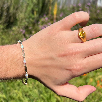 Sunlit view of hand wearing ring on finger and 7.25 inch-long lab-created opal tennis bracelet on wrist against blurred natural background.