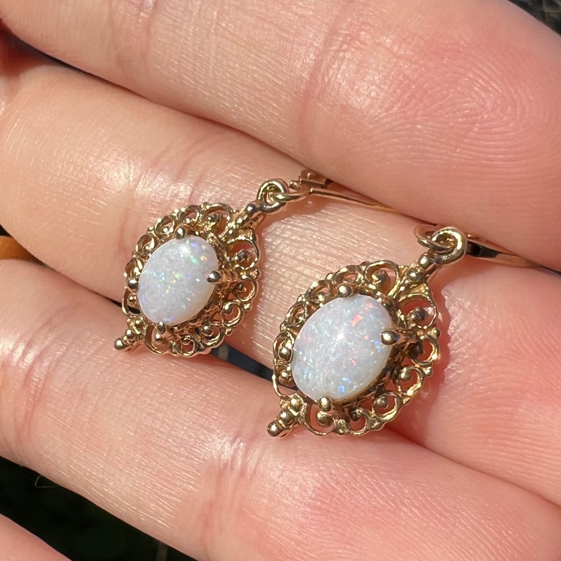 A pair of vintage yellow gold Australian light opal dangle earrings shown close-up in the sunlight, held in a hand and shown from an angle.