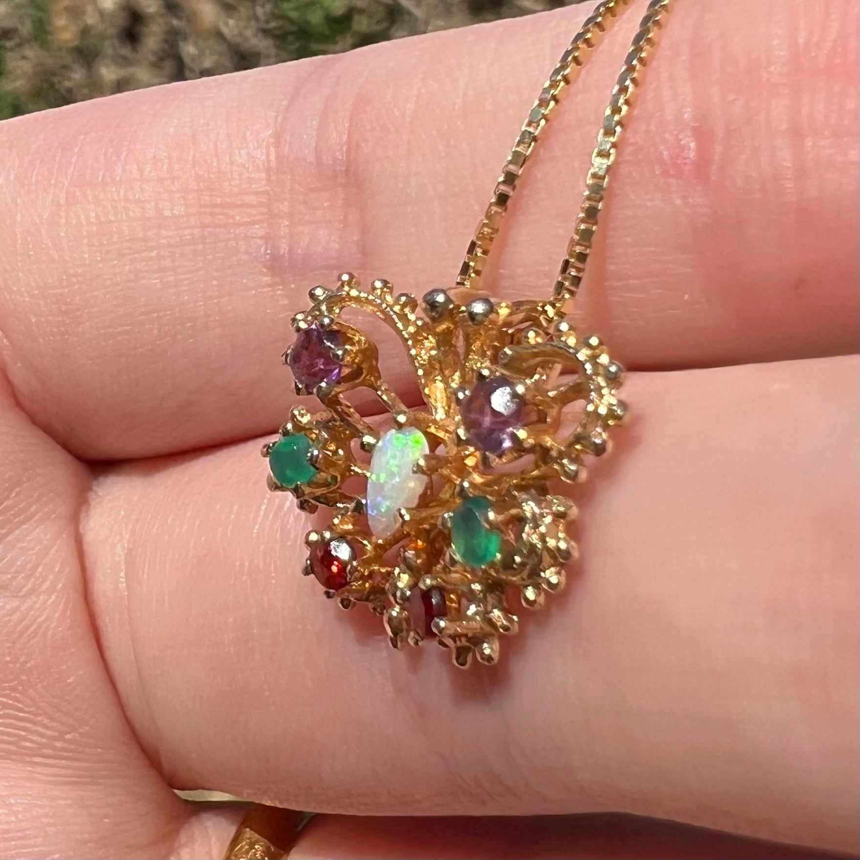 A vintage Australian opal and multi-gemstone butterfly pendant in 14k yellow gold shown up close from angle in sunlight, resting on fingers.