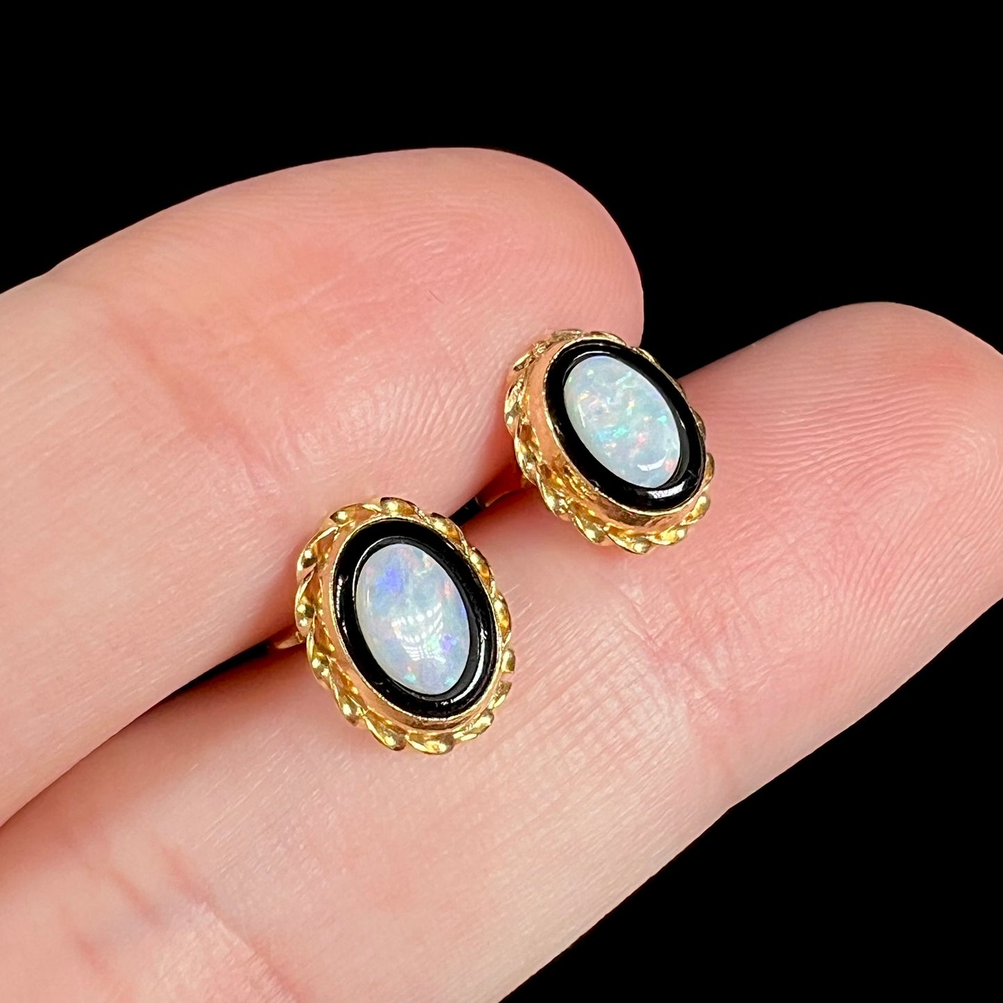 Angled close-up of a pair of yellow gold vintage Australian opal in onyx stud earrings held between two fingers on black background.