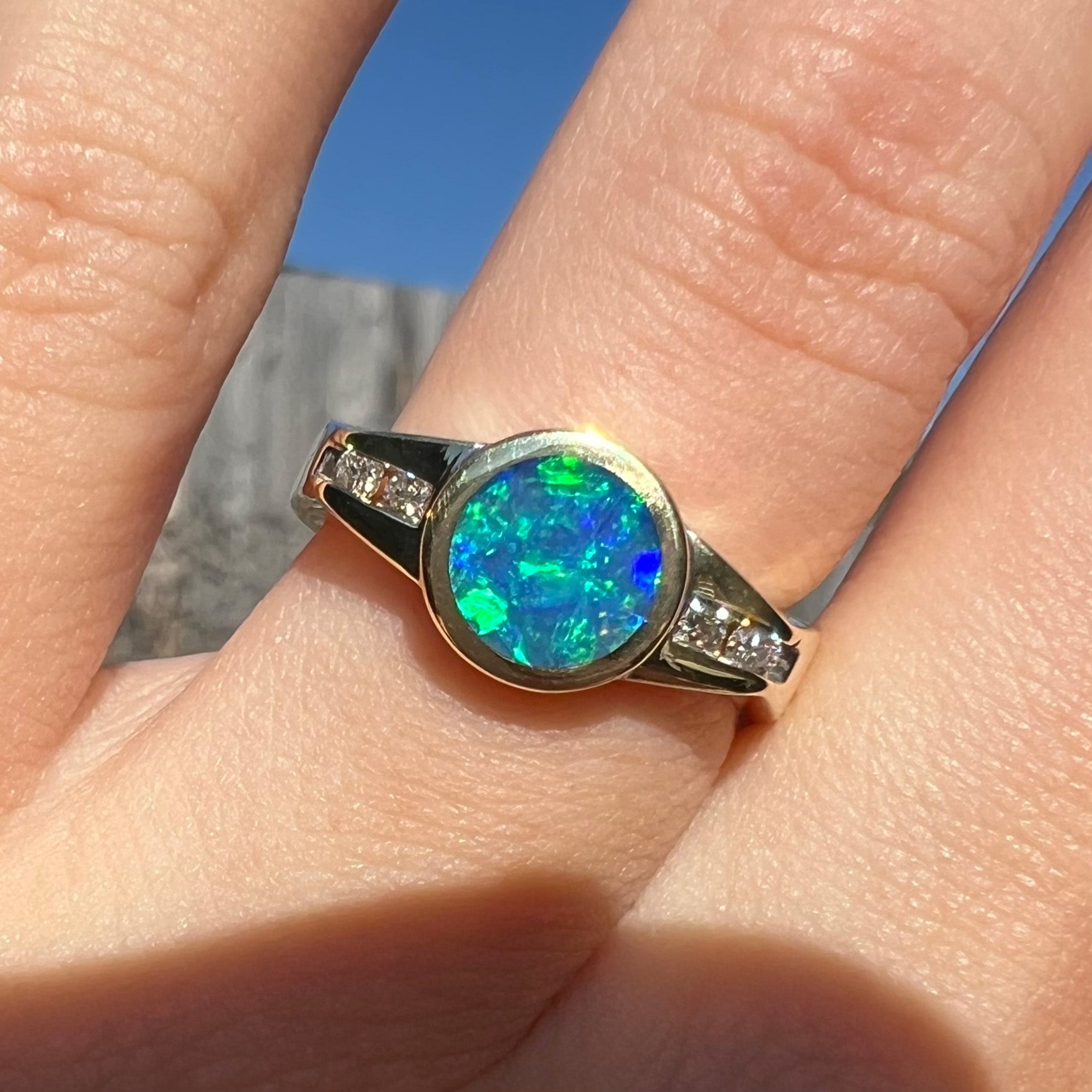 Close-up of a vintage boulder opal engagement ring in yellow gold with diamond accents by Luciano Bortone, shown on a finger in the sunlight.