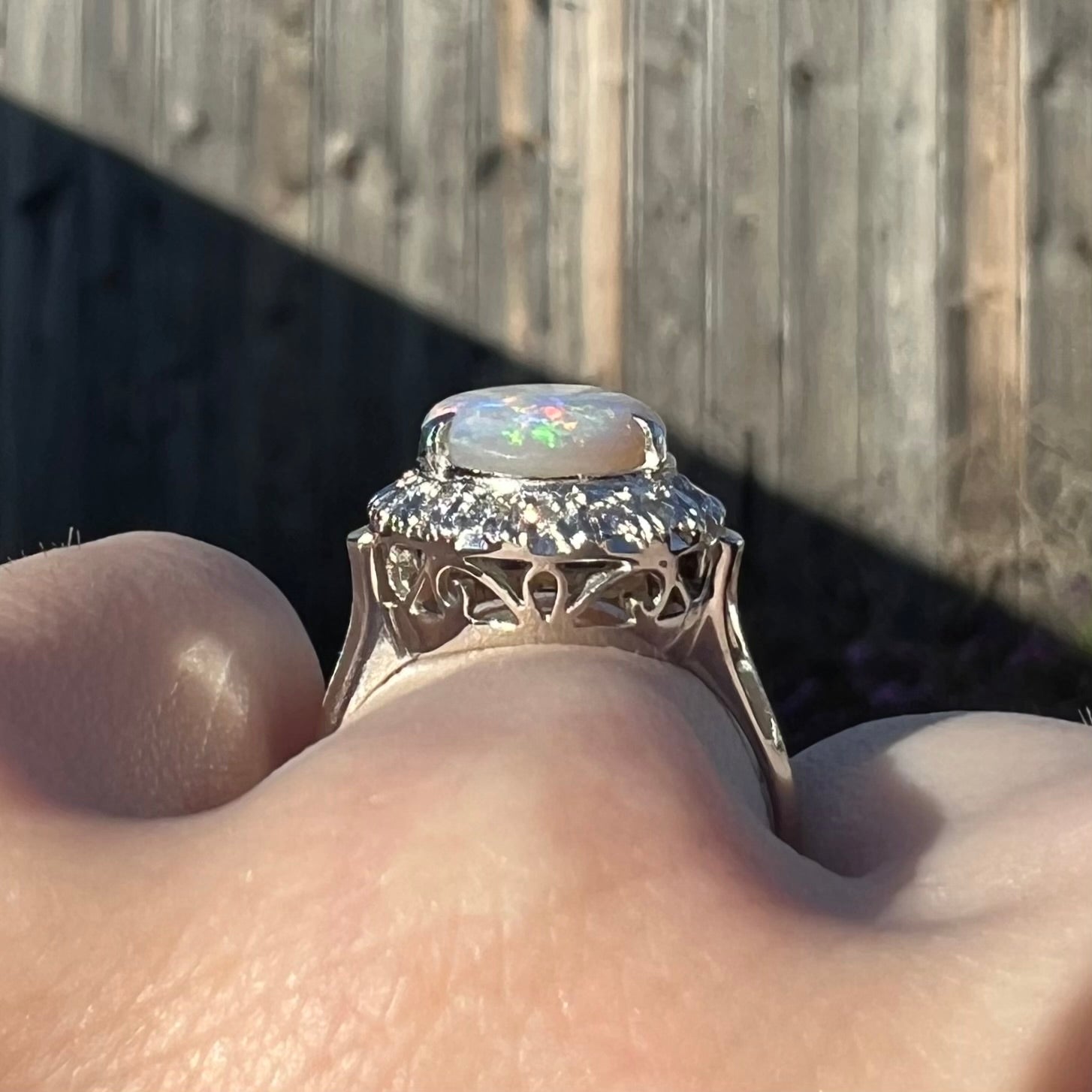 A filigree opal and diamond halo enggagement ring on a hand in the sunlight.