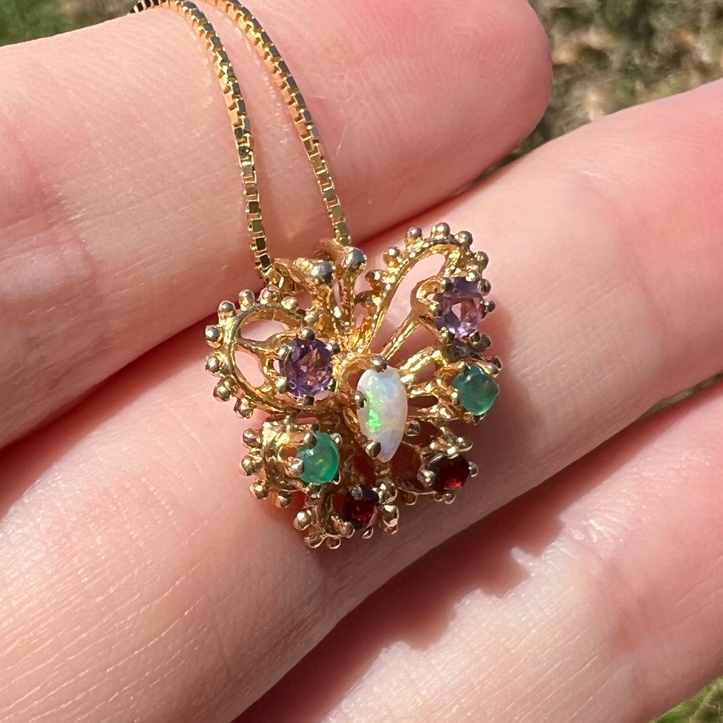Angled close-up of a vintage gear design gold butterfly pendant set with different gemstones, resting on top of fingers in sunlight.