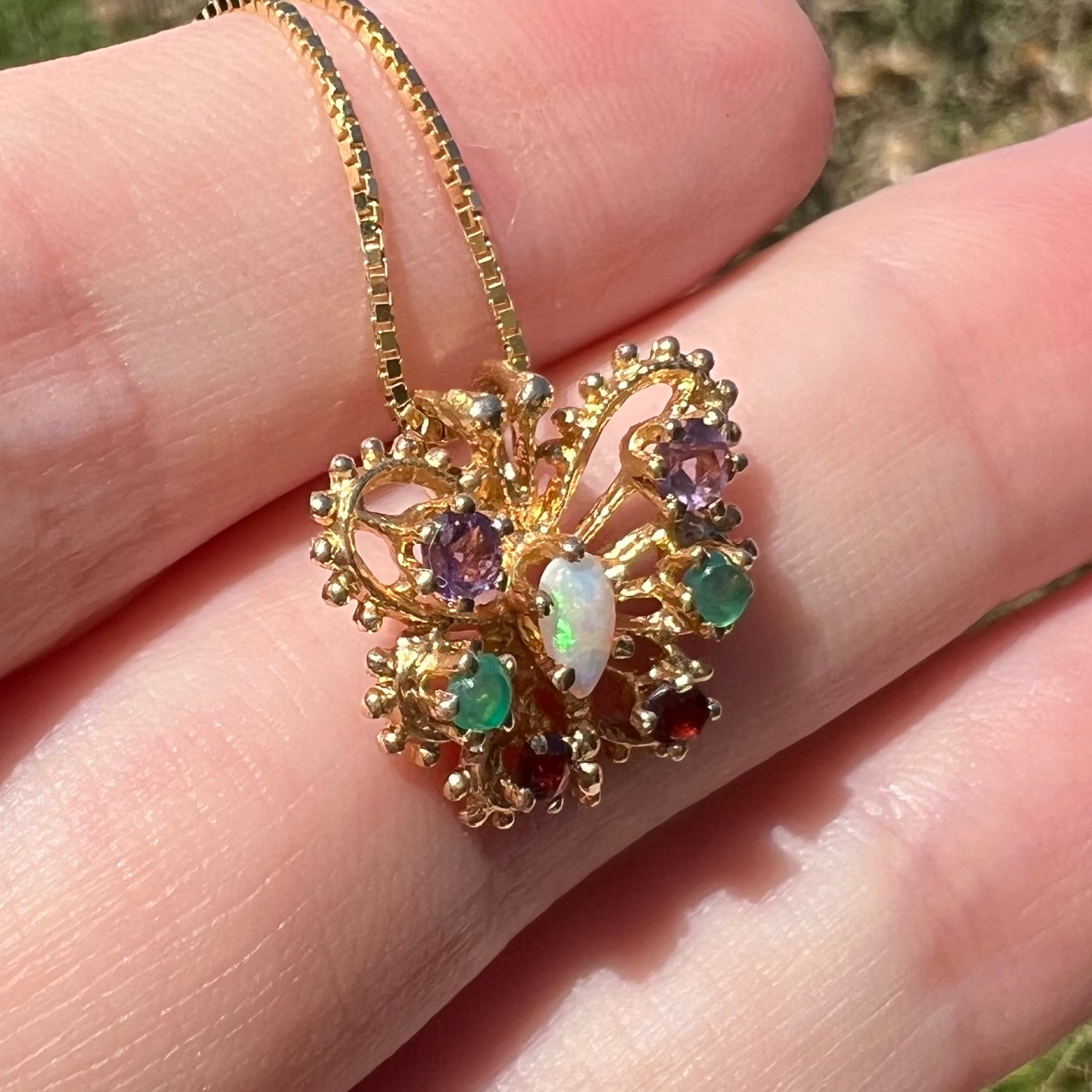 Angled close-up of a vintage gear design gold butterfly pendant set with different gemstones, resting on top of fingers in sunlight.