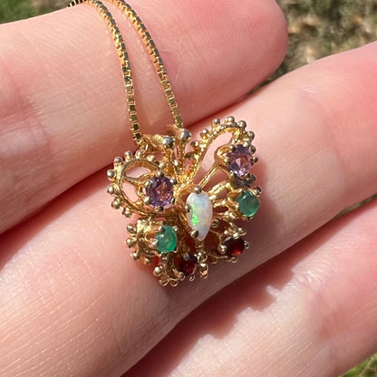 Angled close-up of a vintage gear design gold butterfly pendant set with different gemstones, resting on top of fingers in sunlight.