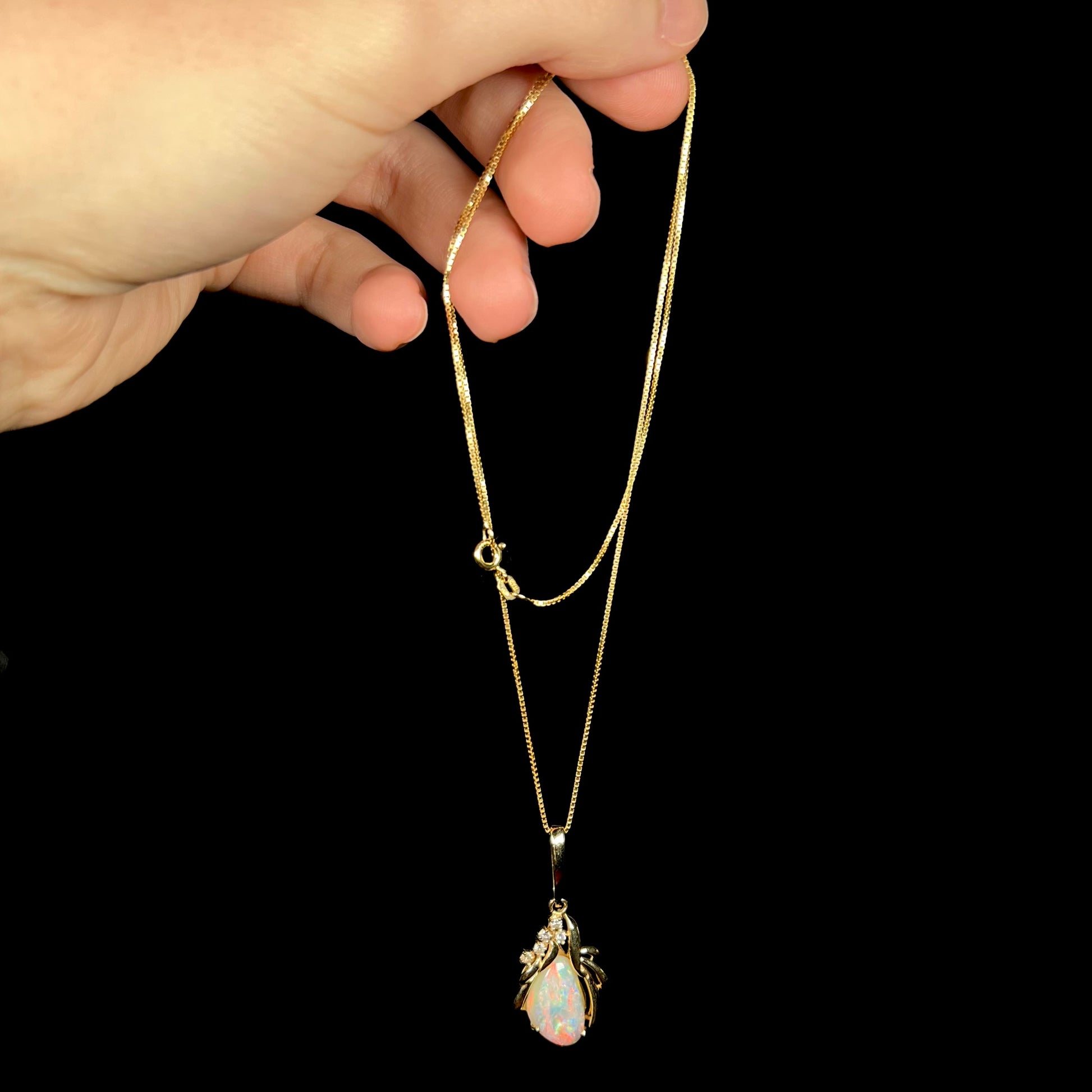 Hand dangling vintage 14k gold Australian opal necklace from box-style chain against black backdrop.