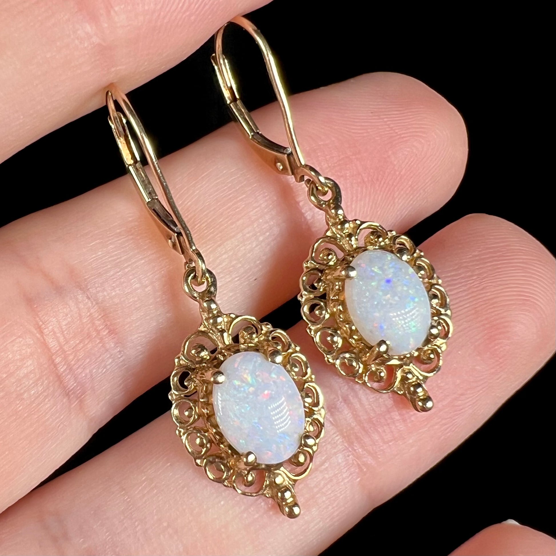 A pair of vintage yellow gold Australian light opal dangle earrings shown close-up in a hand from an angle.
