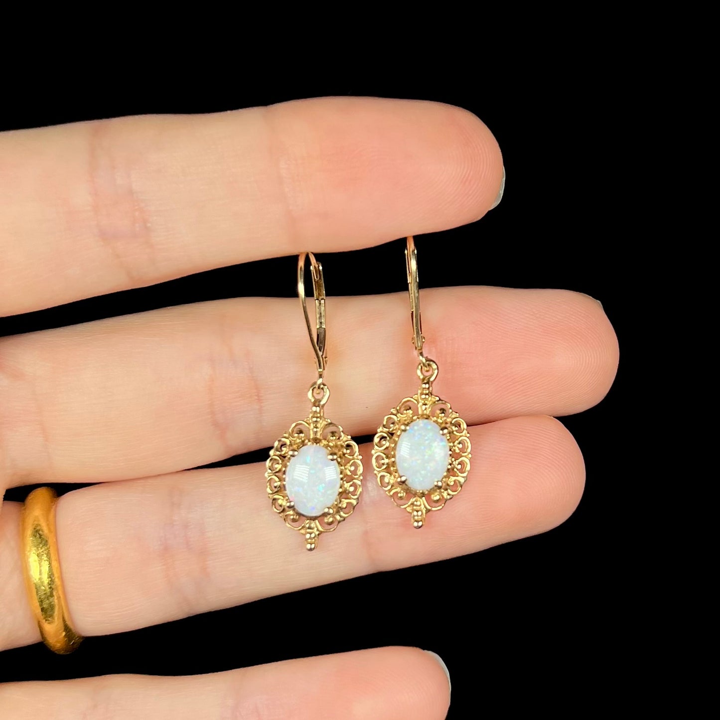 A pair of vintage yellow gold Australian light opal dangle earrings shown in a hand against a black background.