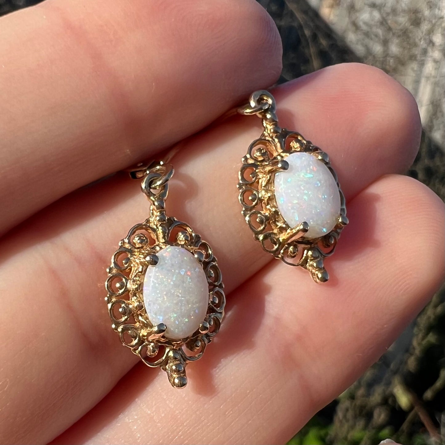Close-up angled sunlit view of a pair of vintage yellow gold Australian light opal dangle earrings shown in a hand.