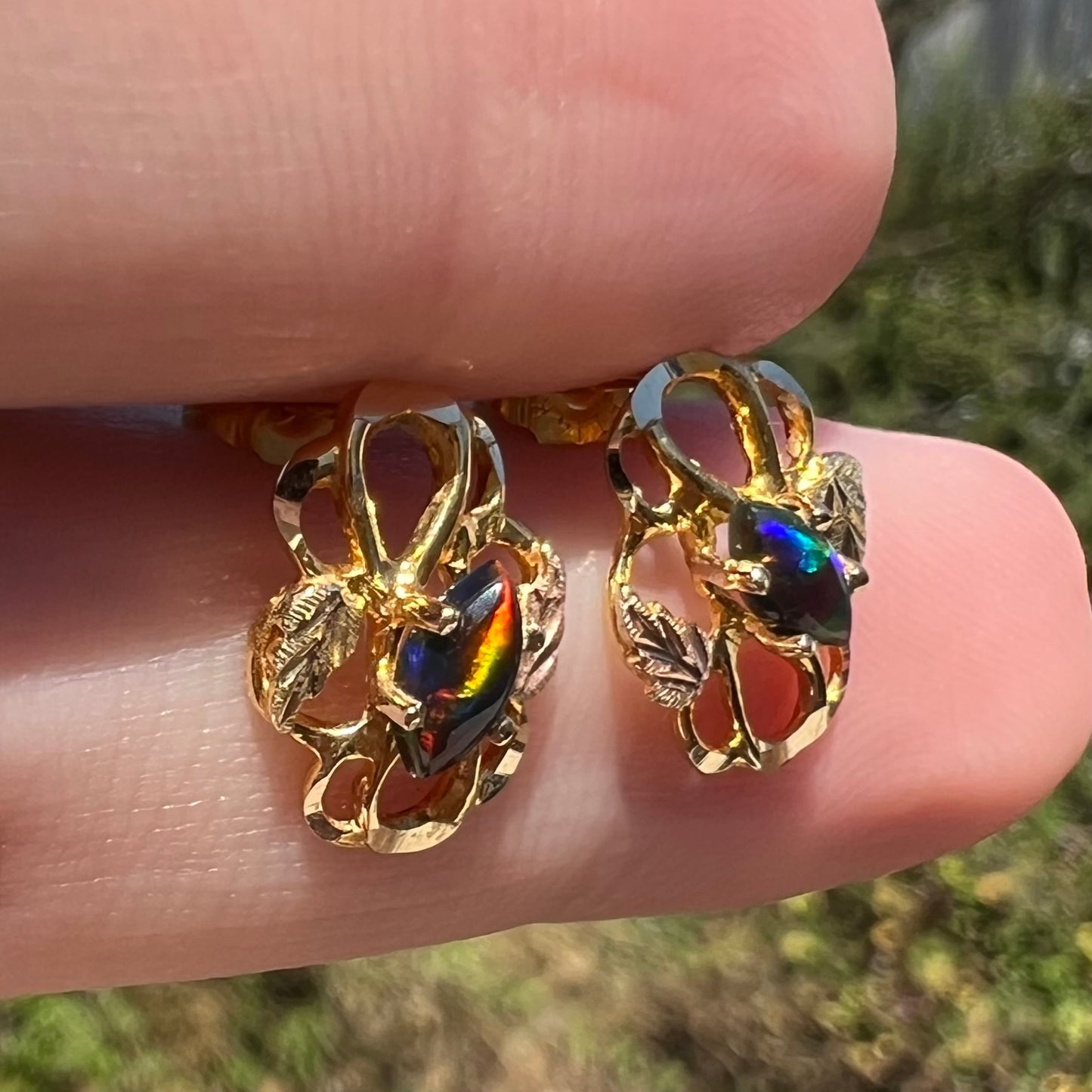 Pair of vintage marquise cut black opal stud earrings with Black Hills gold grape leaf design, held between fingers in sunlight
