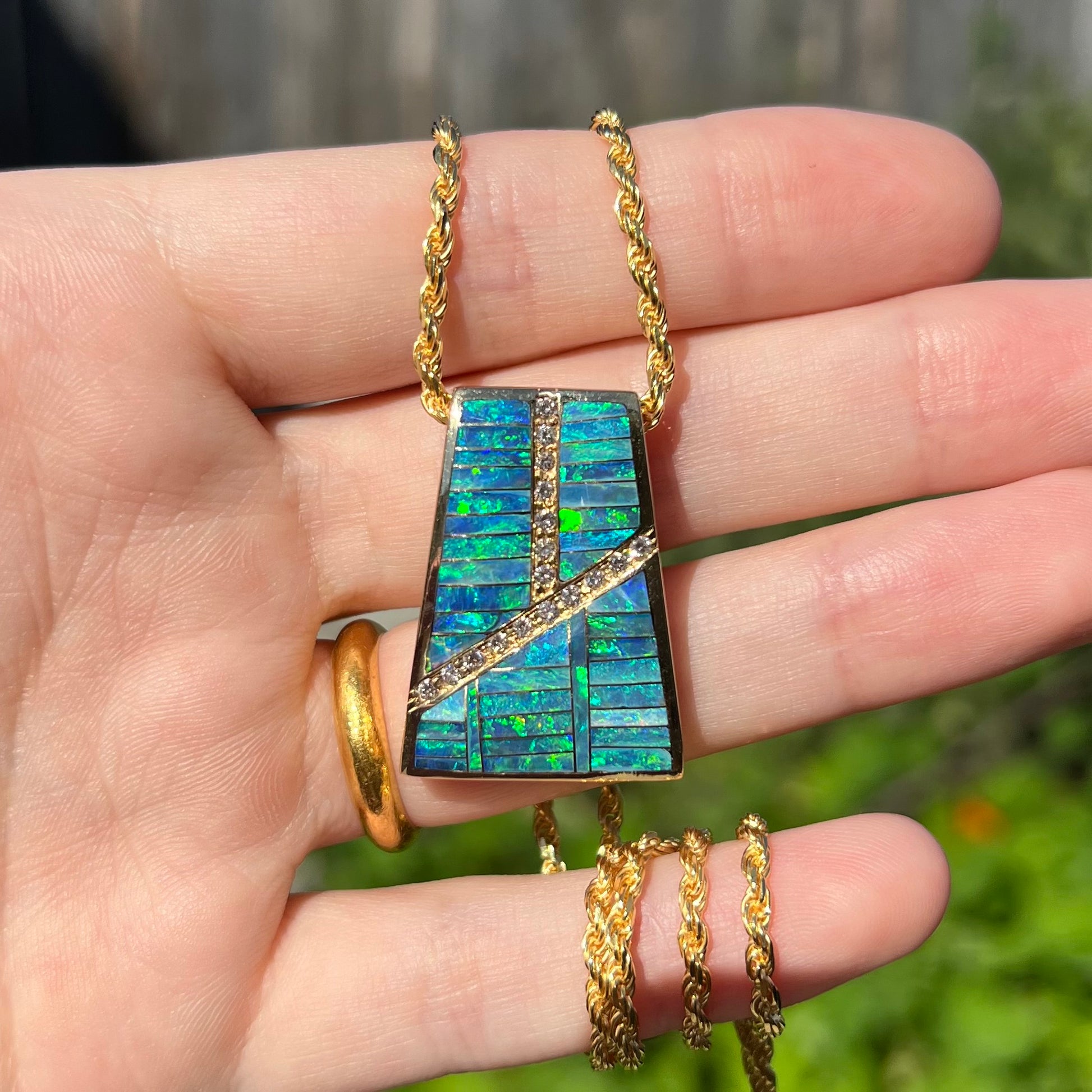 Hand holding a vintage men's opal inlay pendant with diamond accents in the sunlight against a blurred outdoor backdrop.
