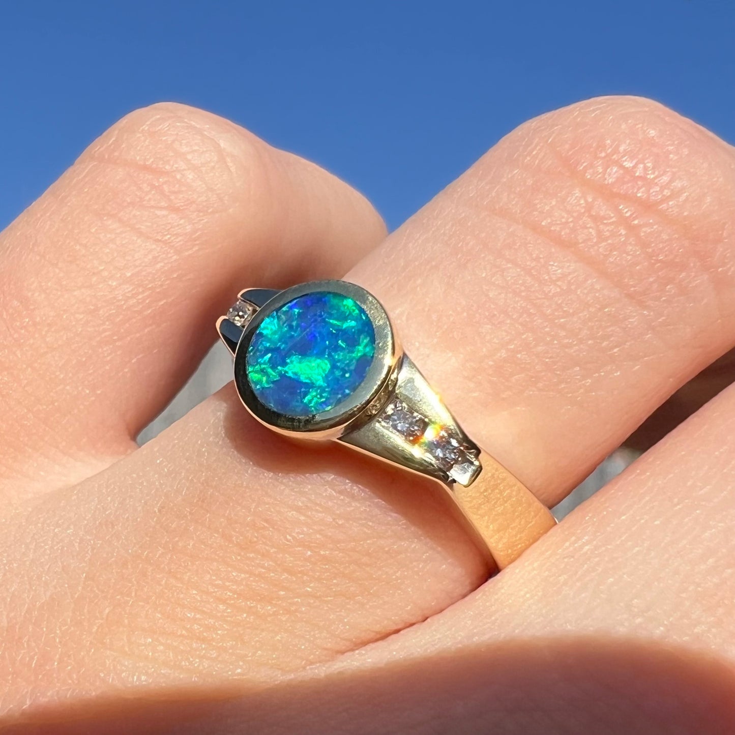 A vintage ring featuring an N4 black boulder opal and diamond accents, worn on a finger and shown from an angle.  The opal shines with green flashes in the sunlight.