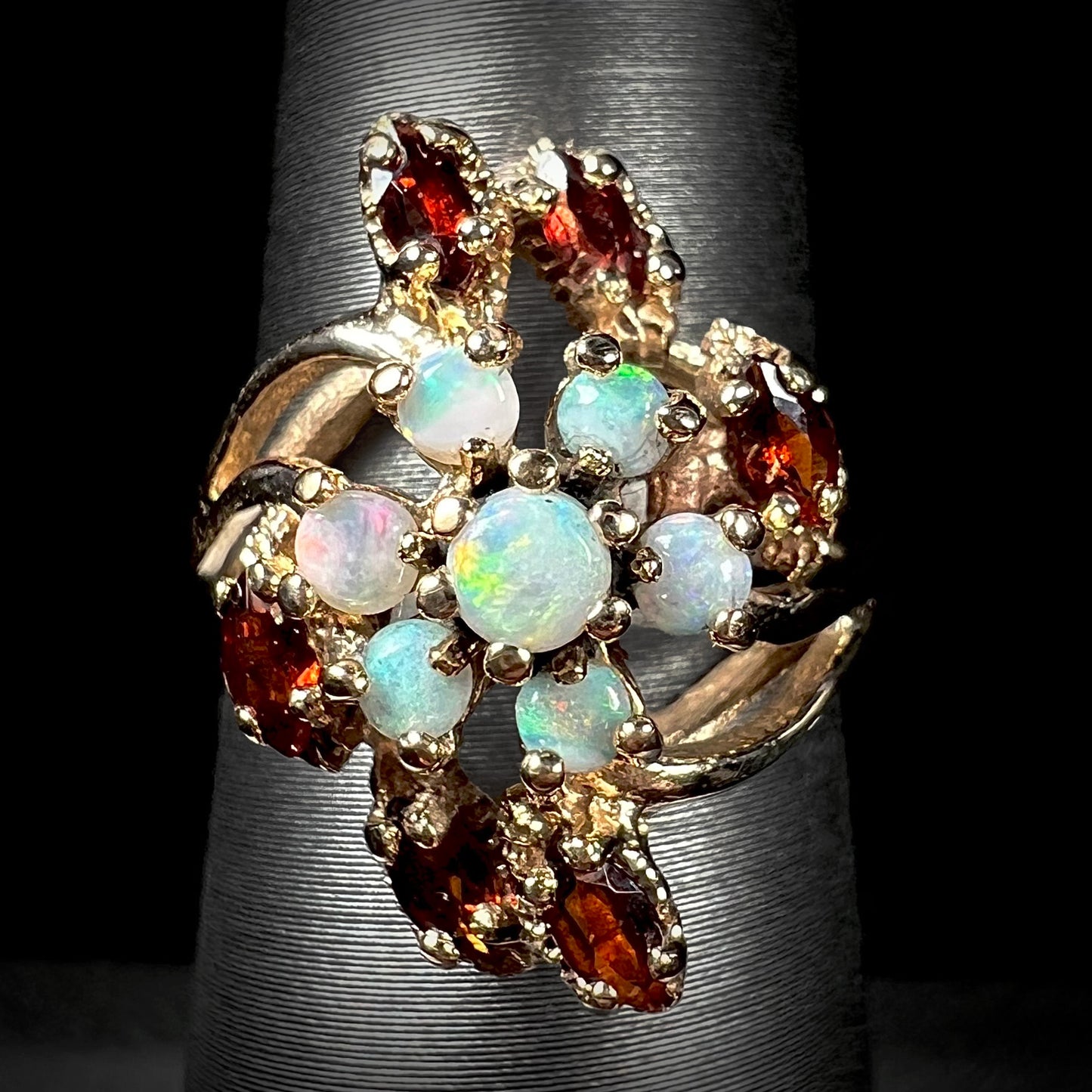 A macro view of a vintage opal and garnet cluster ring in 10k gold on a ring display against a black background.