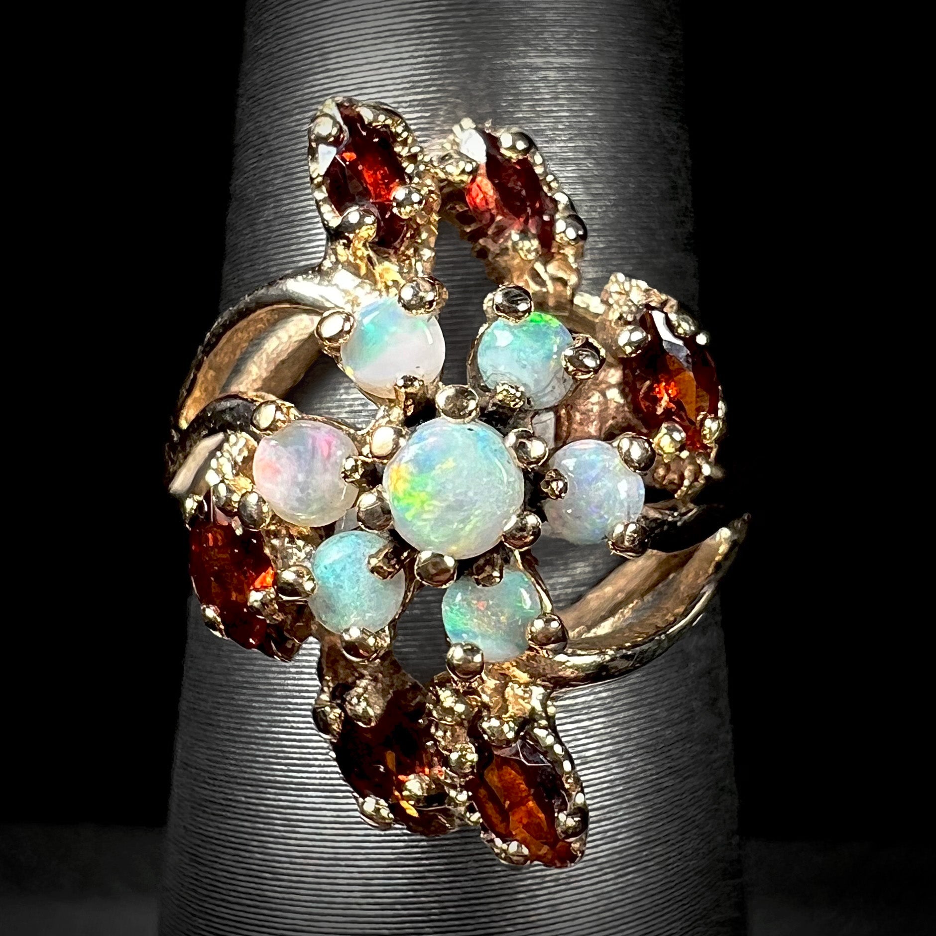 A macro view of a vintage opal and garnet cluster ring in 10k gold on a ring display against a black background.