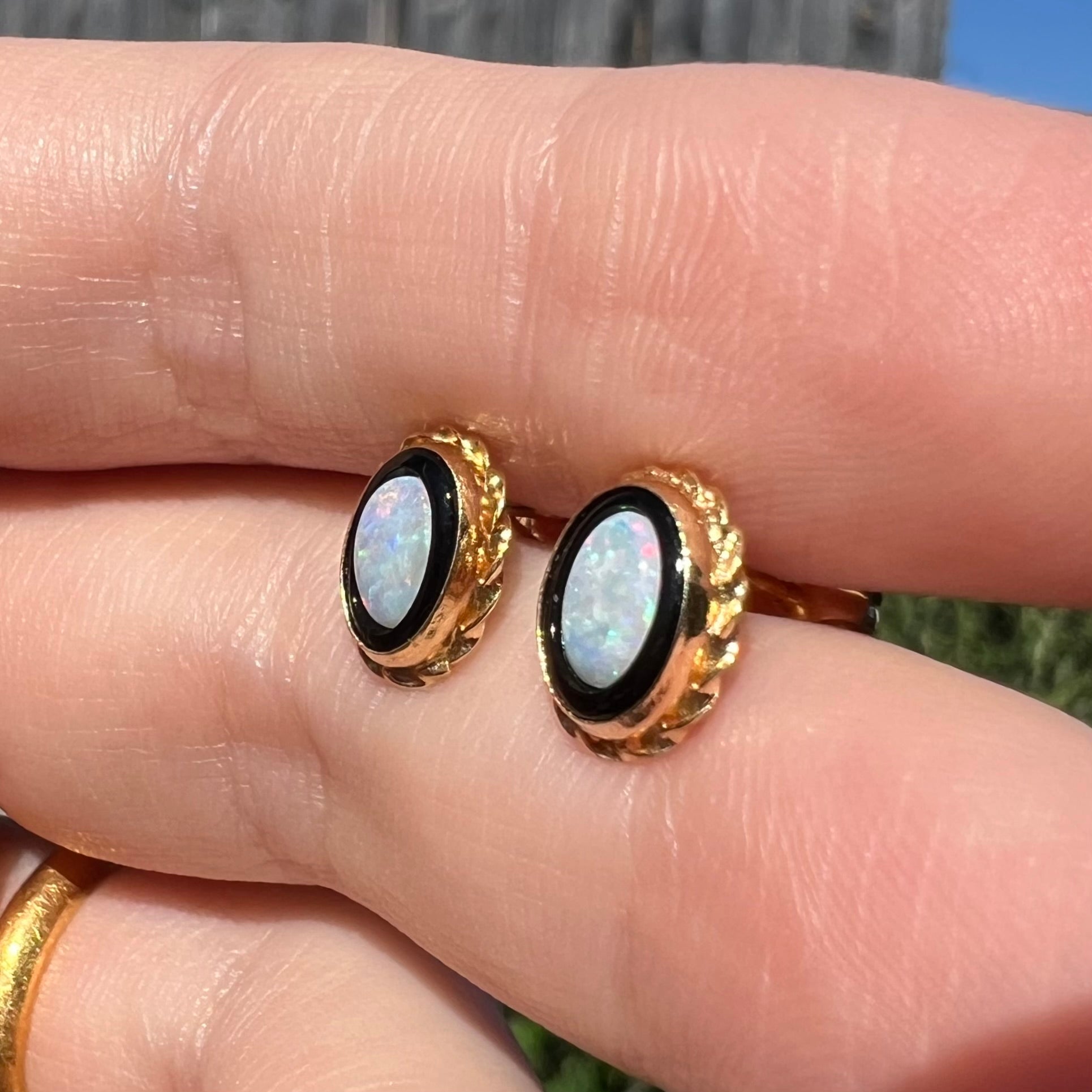 Angled close-up of vintage opal and onyx stud earrings held between two fingers in sunight.