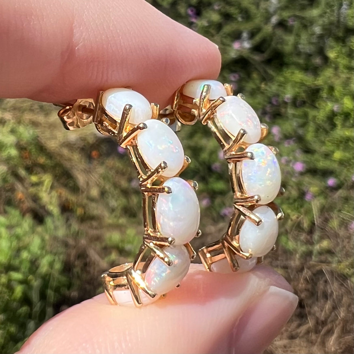 Angled side view close-up of vintage opal semi-hoop earrings 14k gold, held between two fingers in sunlight