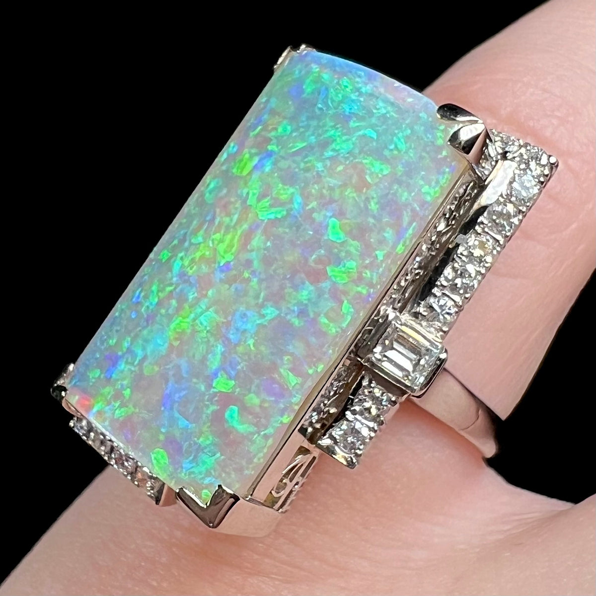 Modified emerald cut diamond accent set amid row of round brilliant cut diamond accents visible along side edges of vintage platinum opal cocktail ring, worn on pinky finger.