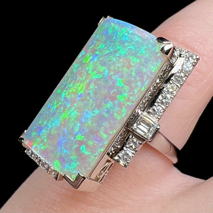 Modified emerald cut diamond accent set amid row of round brilliant cut diamond accents visible along side edges of vintage platinum opal cocktail ring, worn on pinky finger.