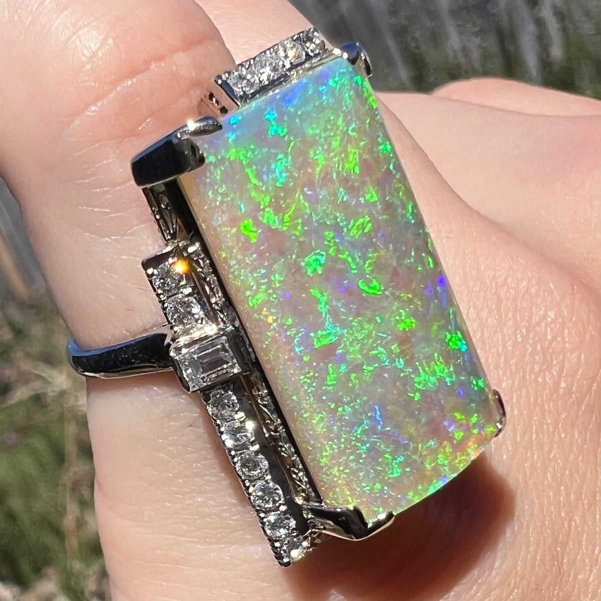 A row of diamonds is seen sparkling in the sunlight, lining the side of a vintage, rectangle-cut opal ring in Japanese platinum, worn on finger.