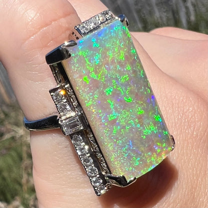 A row of diamonds is seen sparkling in the sunlight, lining the side of a vintage, rectangle-cut opal ring in Japanese platinum, worn on finger.