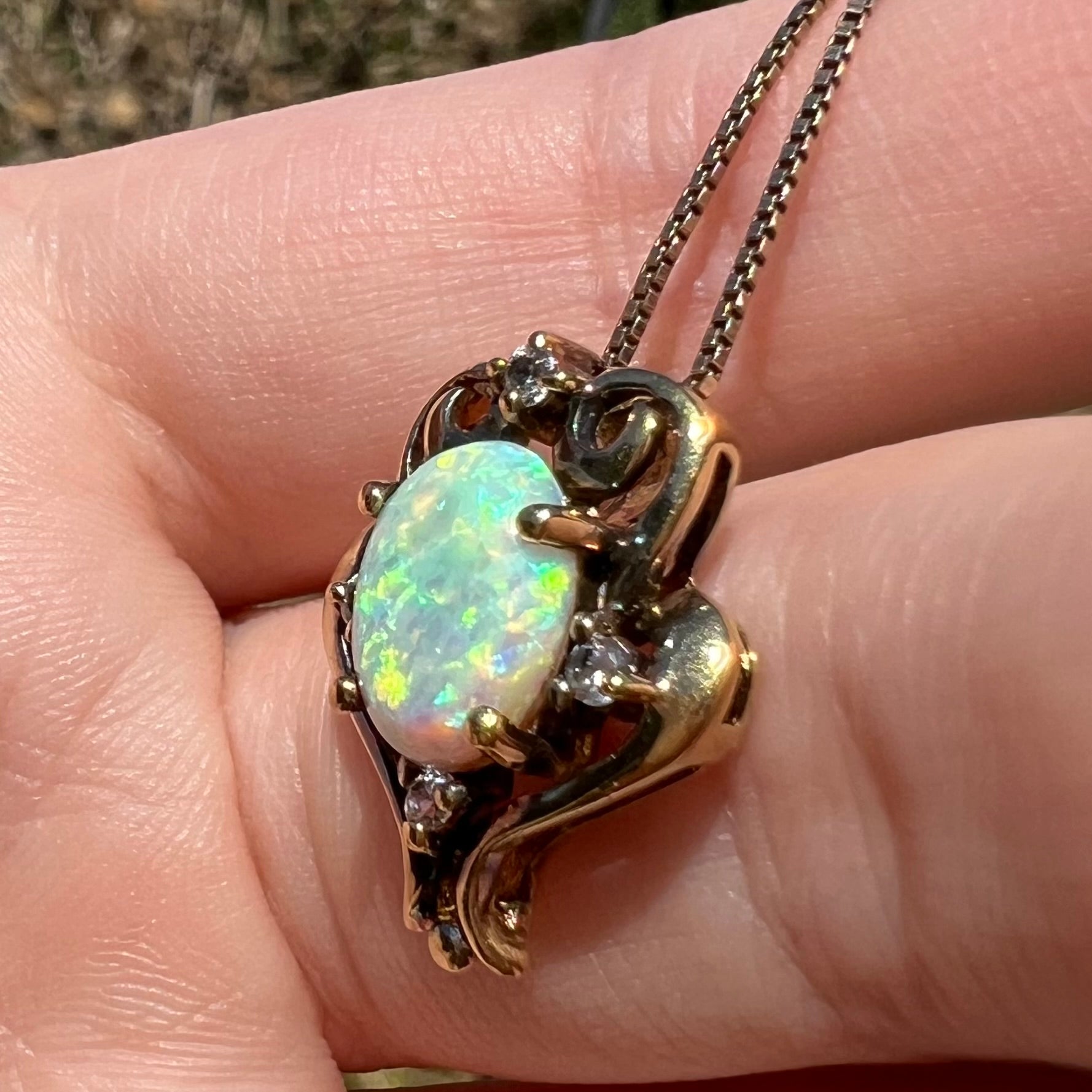 A vintage synthetic opal and CZ pendant with an aged patina over gold-tone metal, shown from an angle close-up in sunlight.