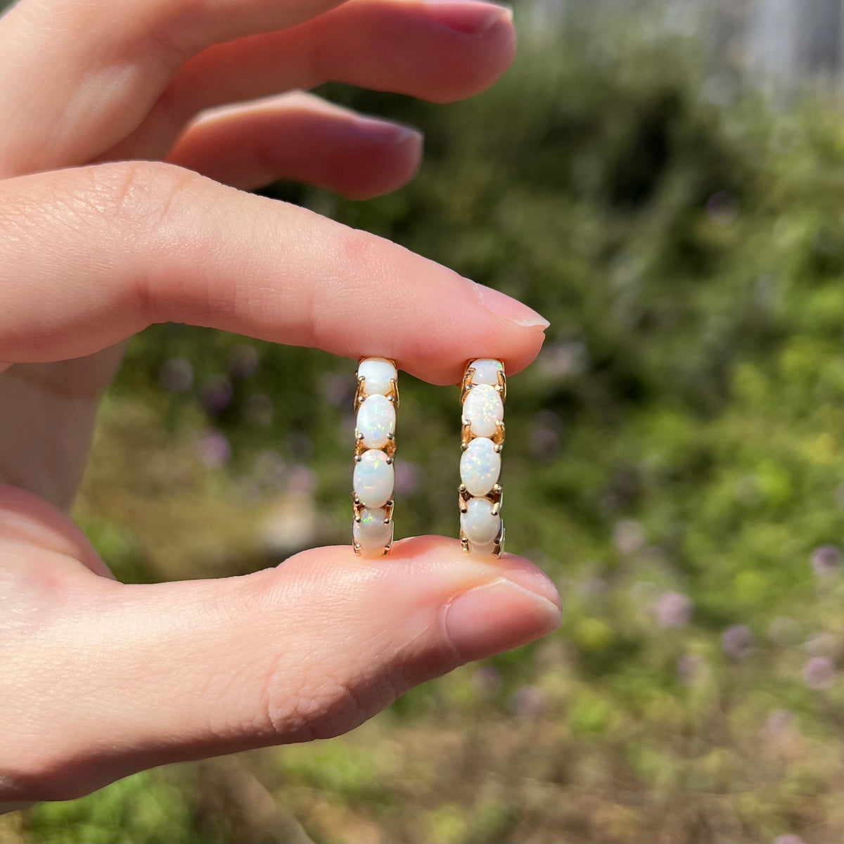 Hand holding pair of vintage gold women's opal hoop earrings between two fingers in sunlight