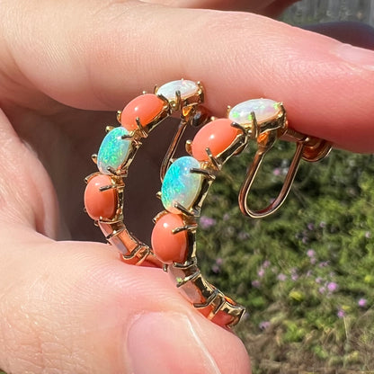 Pair of vintage c.1990s women's 14k gold opal and coral semi-hoop earrings with non-pierced screw backs, held between two fingers and shown from the side view in sunlight