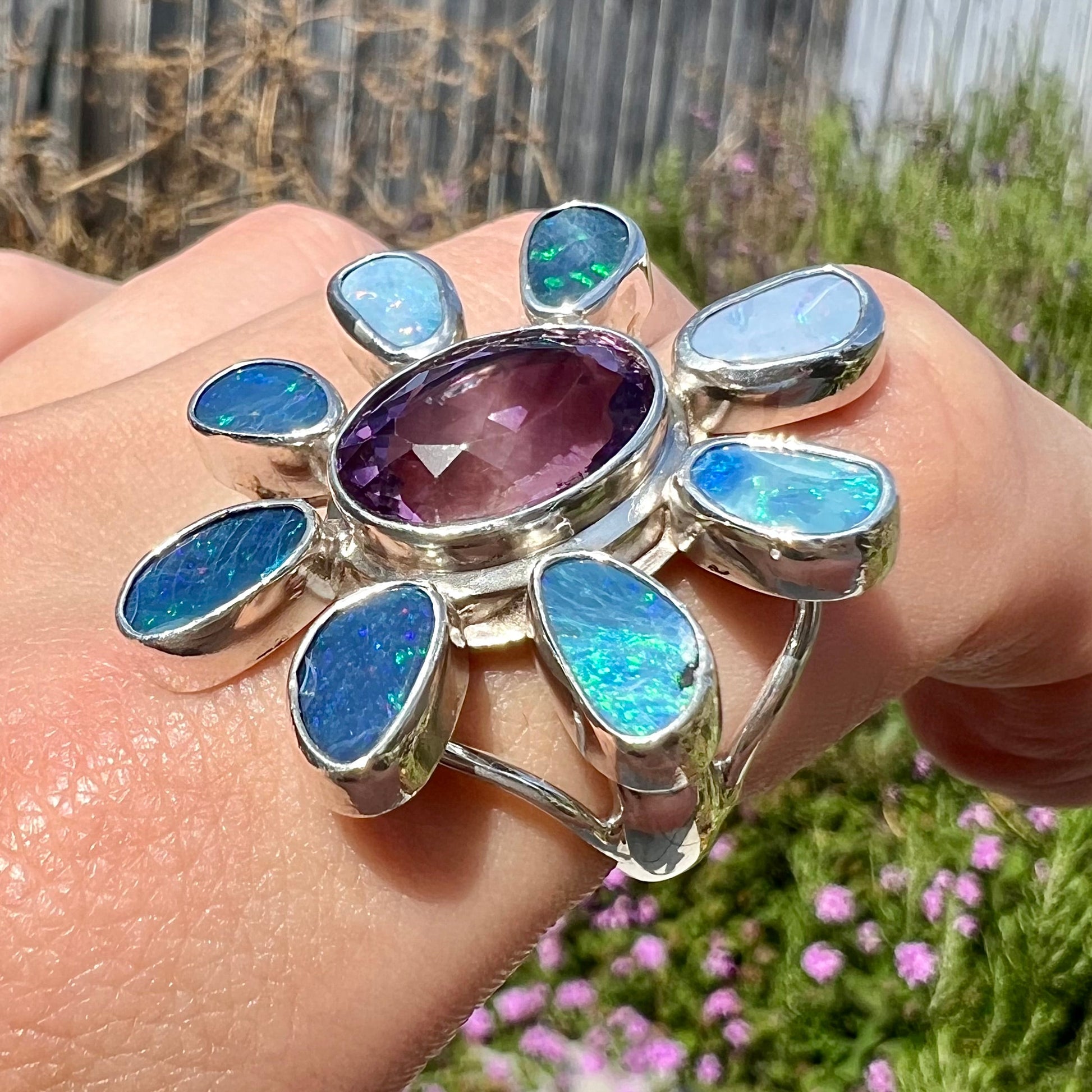 A sterling silver flower shaped ring set with an oval cut amethyst in the center and black opal doublet petals.