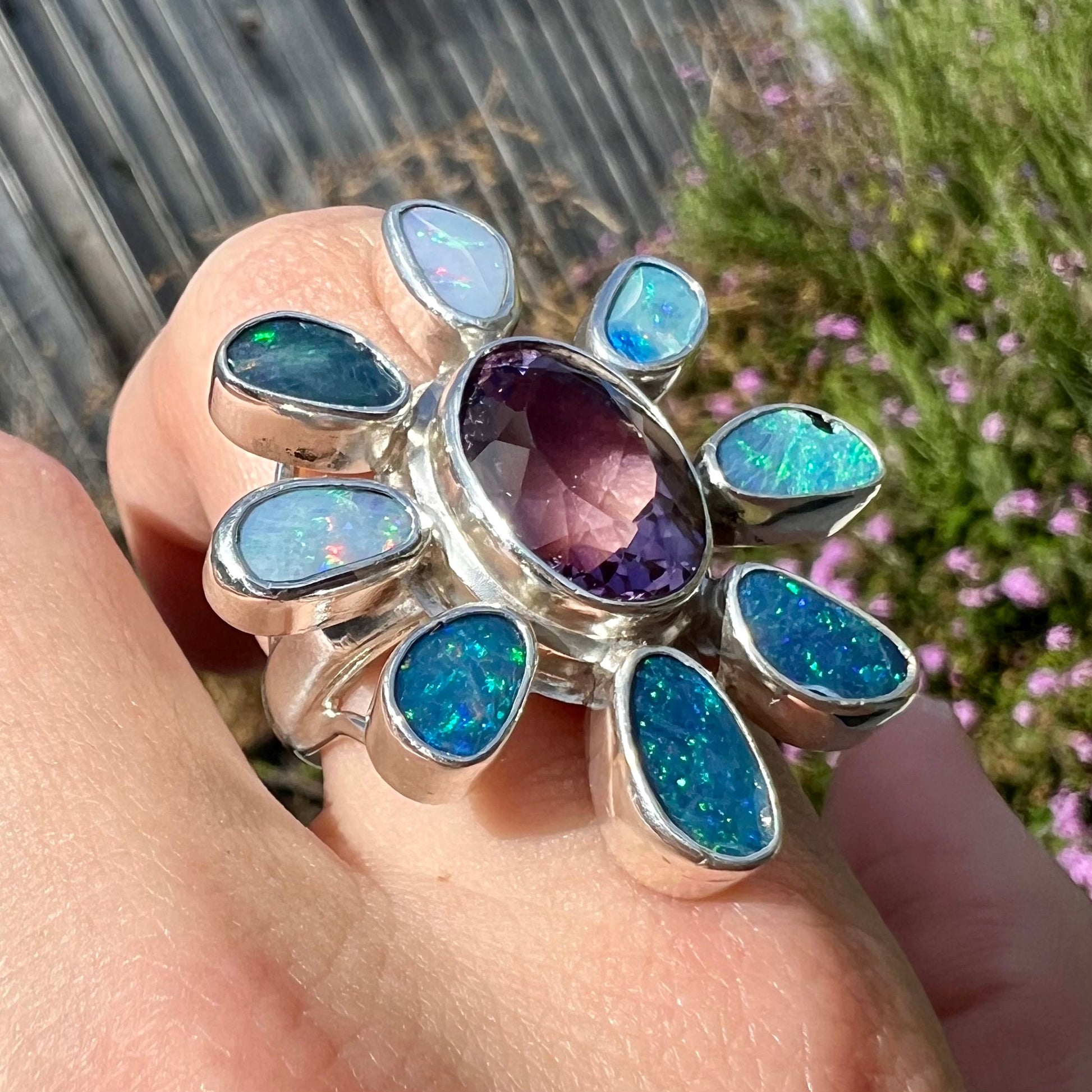A sterling silver flower shaped ring set with an oval cut amethyst in the center and black opal doublet petals.