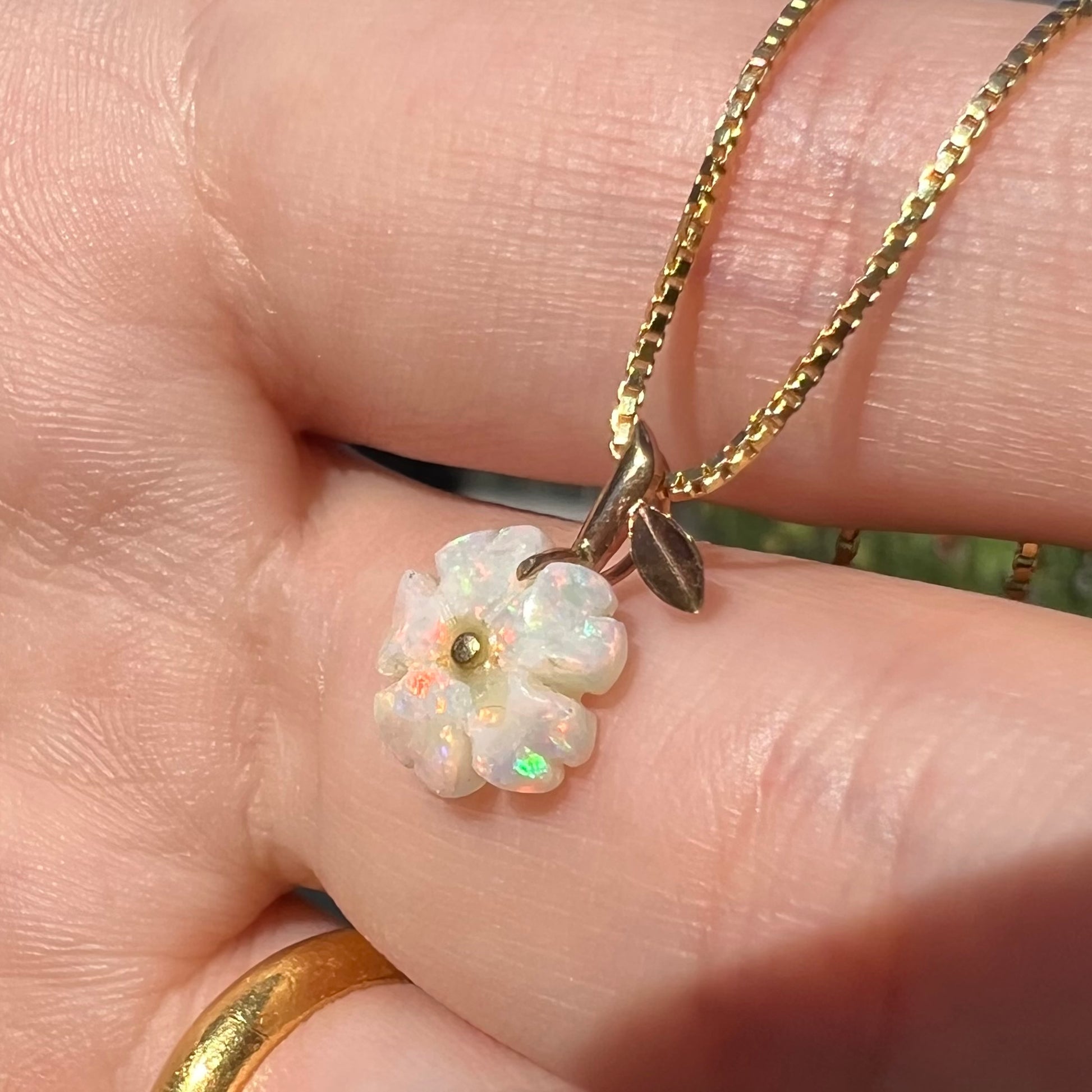 A white Australian opal flower necklace with a yellow gold stem and leaf-design bail shown close-up in a hand from an angle under sunlight.