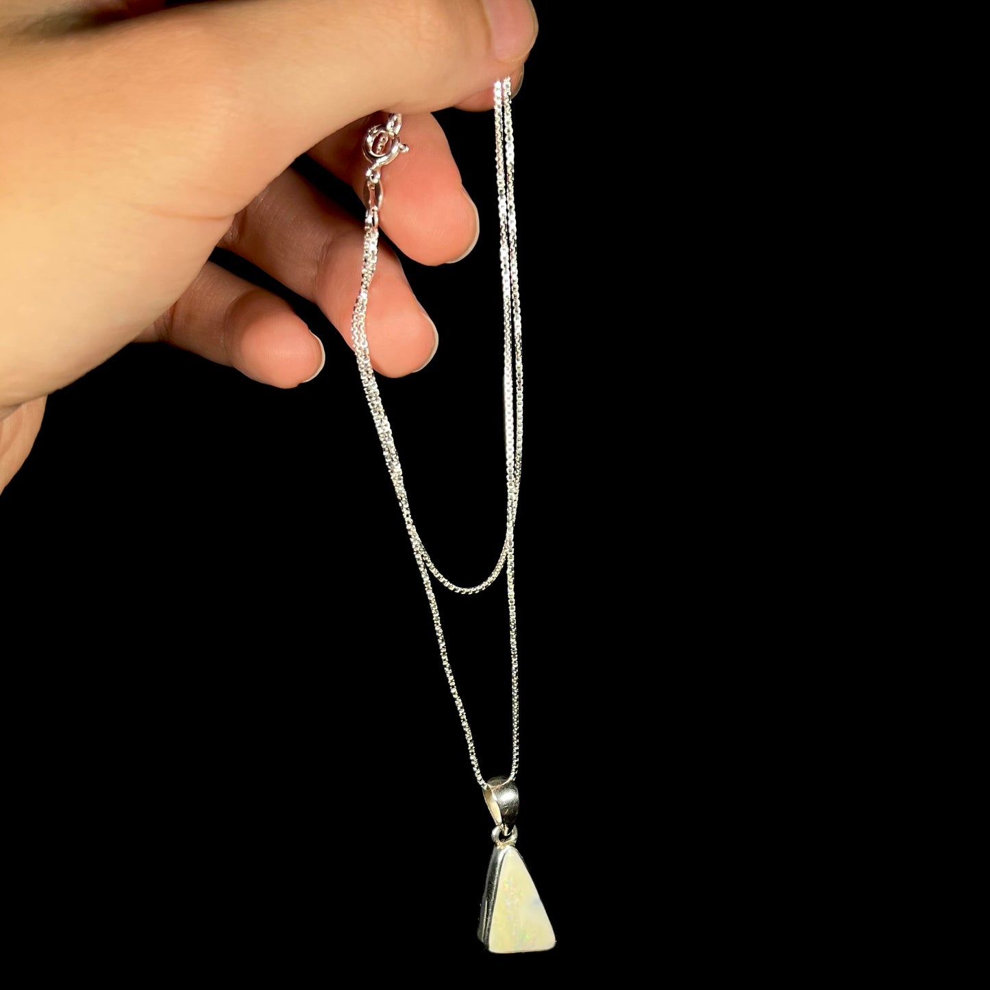 A white Australian boulder opal necklace in .950 silver, shown dangled by a hand from its chain.