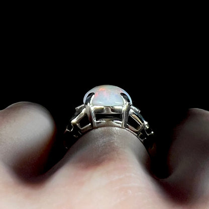 Side gallery profile view of a white gold Australian opal ring, worn on a hand; the ring's prong-set basket and the opal's dome are in focus