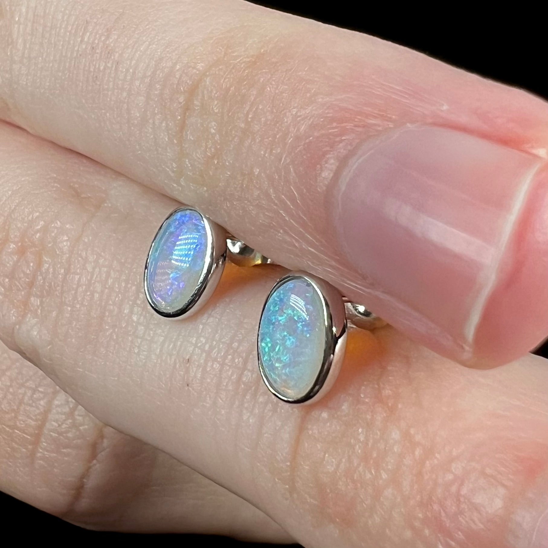 Pair of white gold bezel set Australian opal stud earrings shown close-up from angle, held between fingers.