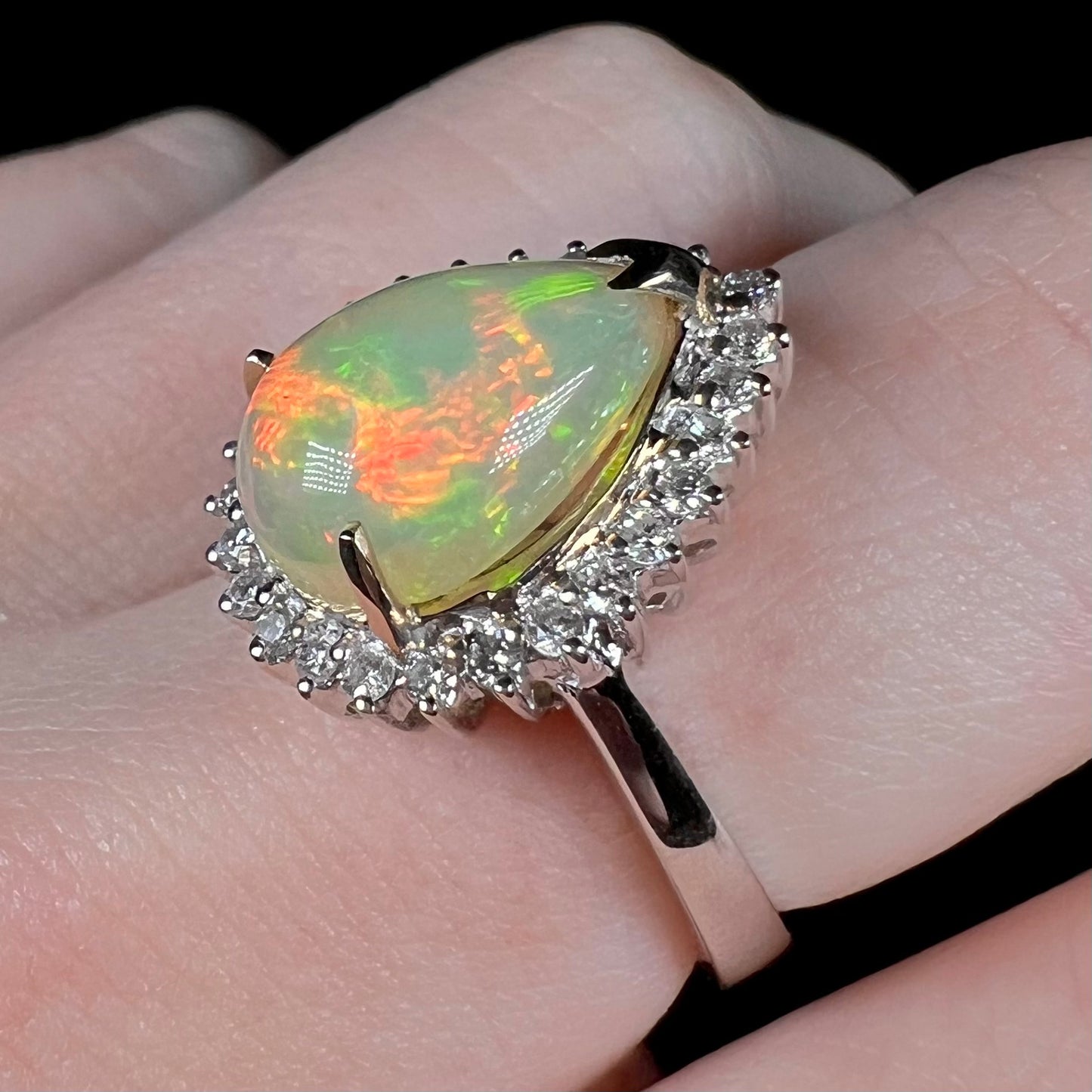 White gold fire opal and diamond halo ring shown close-up on a finger, angled to focus on the ring's single shank design.