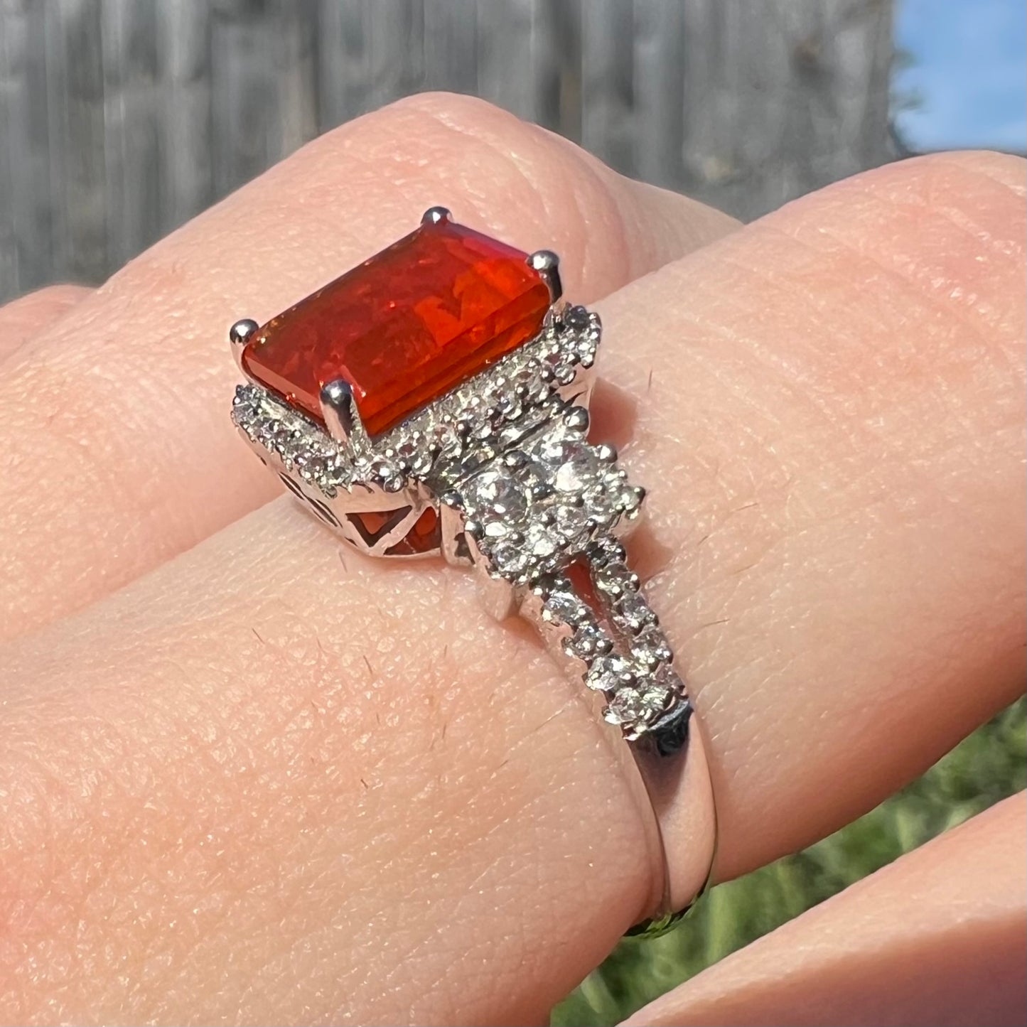 Finger wearing a 14k white gold fire opal and zircon ring shown close-up from an angled view in natural light against blurred outdoor background.