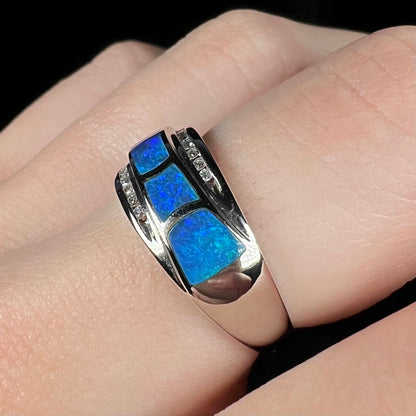 Angled close-up view of white gold ring flush-set with inlaid blue Australian opals and channel-set accent diamonds, worn on finger