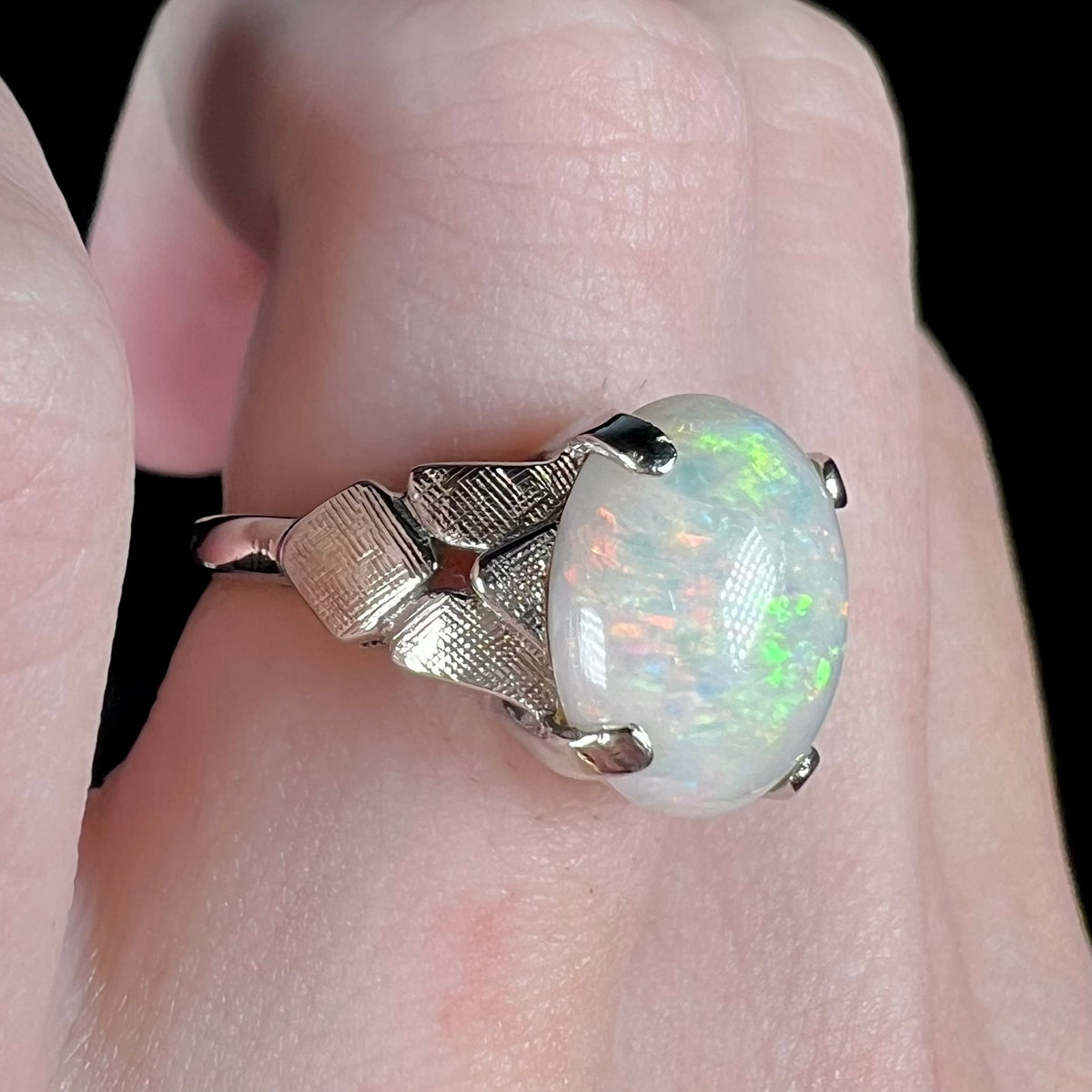 A white gold Australian opal ring, worn on a finger and shown close-up; the opal shows orange and green flashes of color