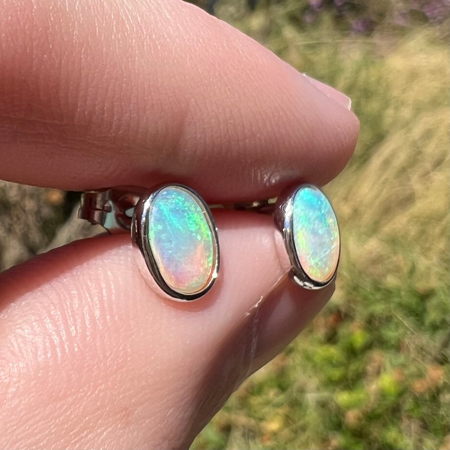 Angled close-up of white gold opal stud earrings shown between two fingers in the sunlight.  The opals have green play of color.