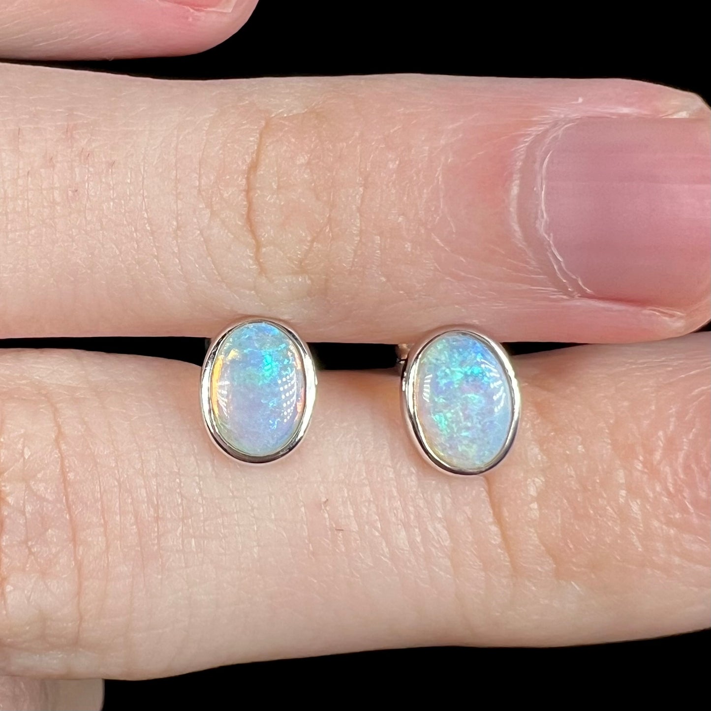 Close-up of pair of 18k white gold oval shaped opal stud earrings with delicate blue play of color, held between two fingers.