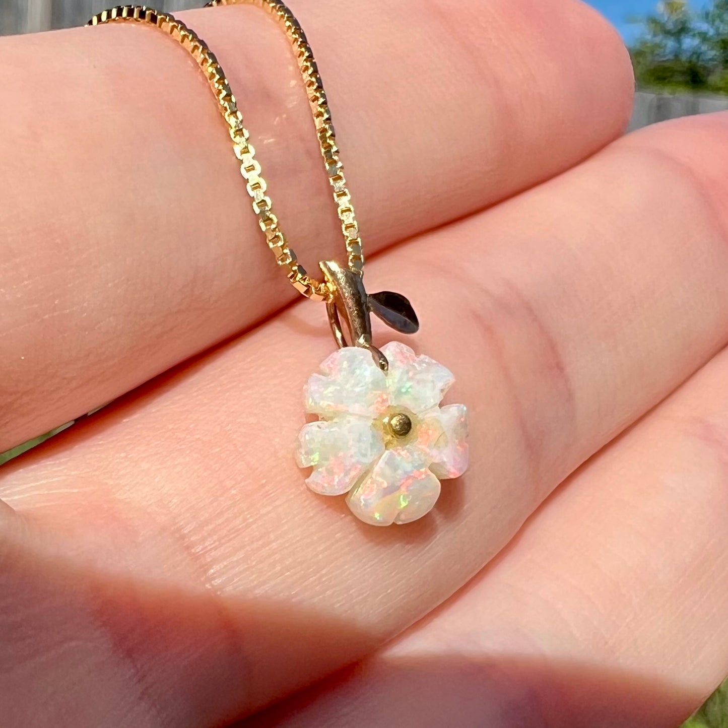 Angled close-up of a white opal flower pendant shown held in a hand under natural light.  The opal has faint red and green play of color.