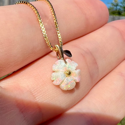 Angled close-up of a white opal flower pendant shown held in a hand under natural light.  The opal has faint red and green play of color.
